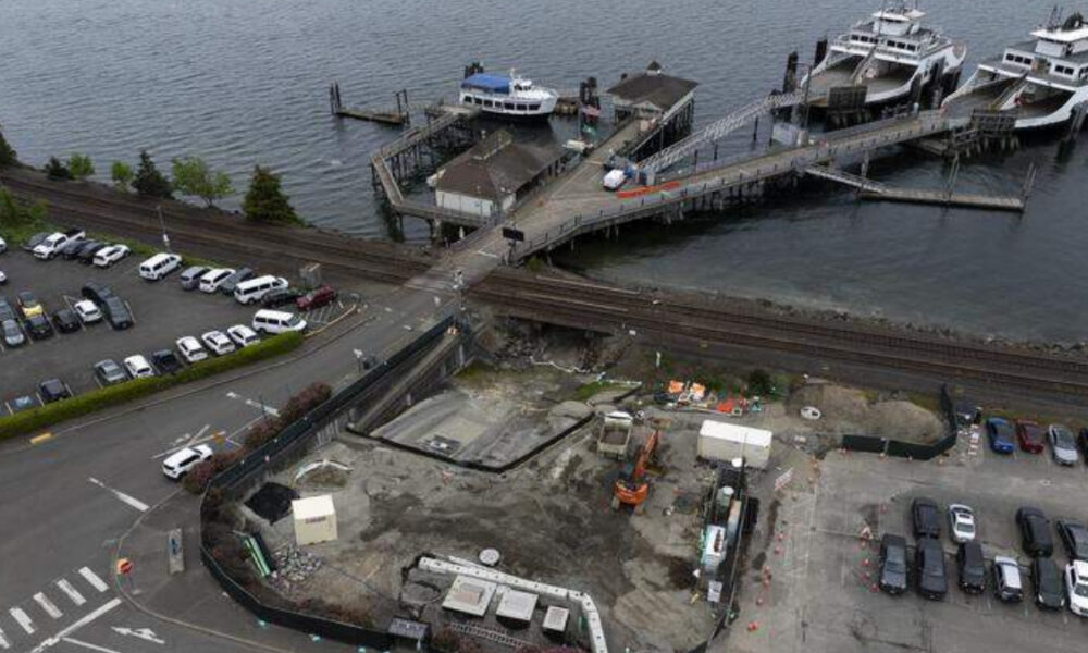 Aerial view of a waterfront construction site with machinery, a pier for boats, nearby parked cars and railroad tracks crossing the area.