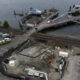 Aerial view of a waterfront construction site with machinery, a pier for boats, nearby parked cars and railroad tracks crossing the area.