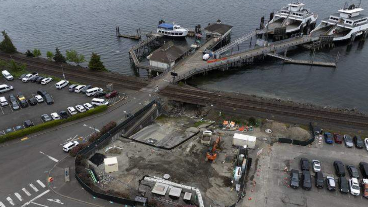 Aerial view of a waterfront construction site with machinery, a pier for boats, nearby parked cars and railroad tracks crossing the area.