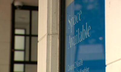 Blue storefront window sign reading 'Space Available' in white cursive text, with names at the bottom of the panel.