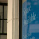 Blue storefront window sign reading 'Space Available' in white cursive text, with names at the bottom of the panel.