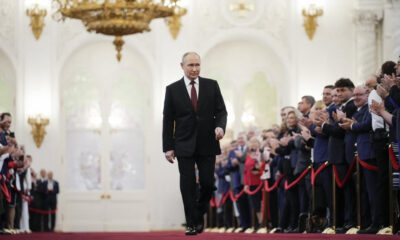 A man in a dark suit walks down a red carpet in a grand, gilded hall as a row of formally dressed people applaud on both sides.