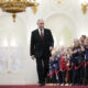 A man in a dark suit walks down a red carpet in a grand, gilded hall as a row of formally dressed people applaud on both sides.