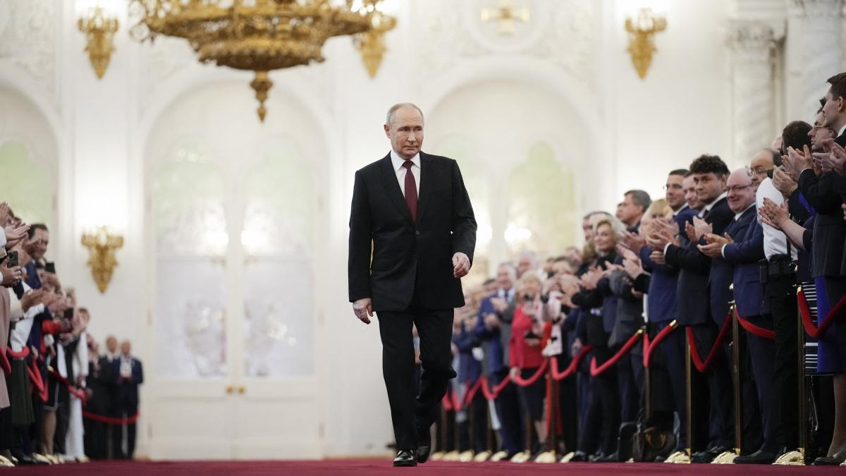 A man in a dark suit walks down a red carpet in a grand, gilded hall as a row of formally dressed people applaud on both sides.