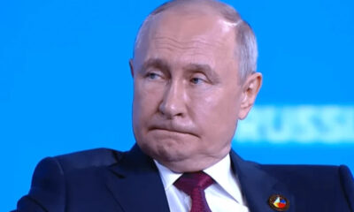 Man in a dark suit and red tie on a bright blue background, looking to the side with a slightly pursed mouth edge. (informative)