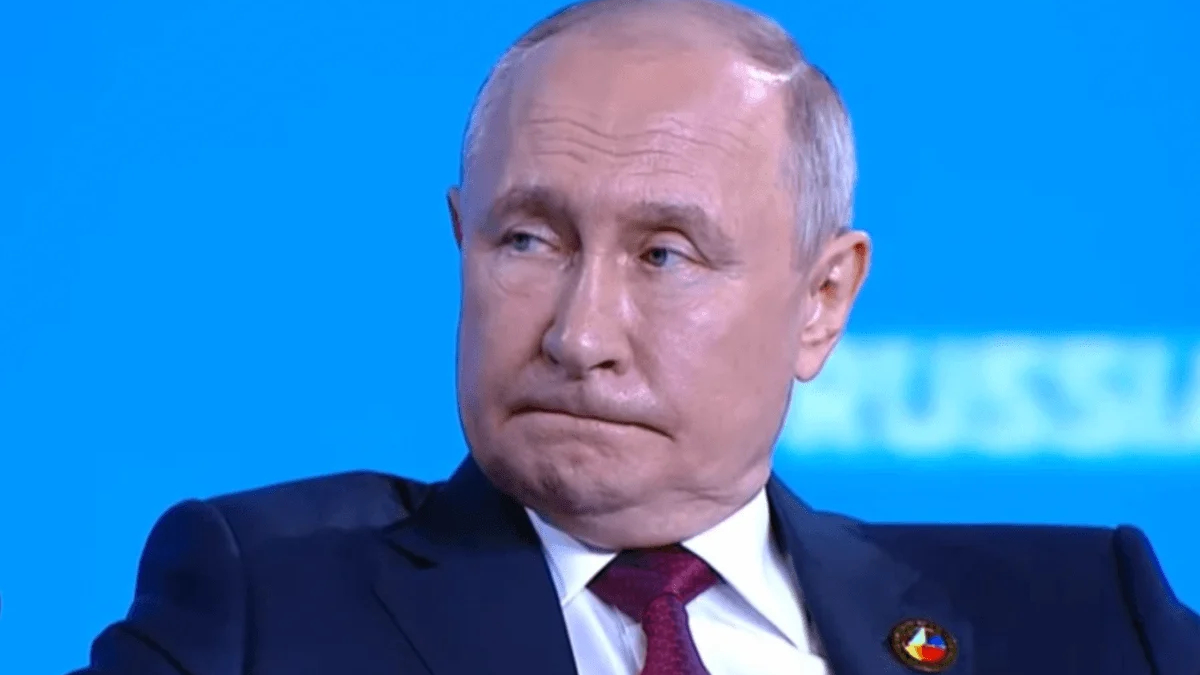 Man in a dark suit and red tie on a bright blue background, looking to the side with a slightly pursed mouth edge. (informative)