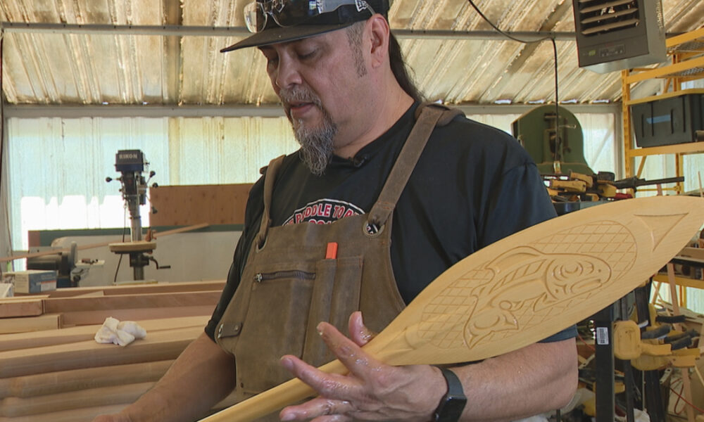 Woodworker in a shop holding a carved wooden surfboard with a fish design