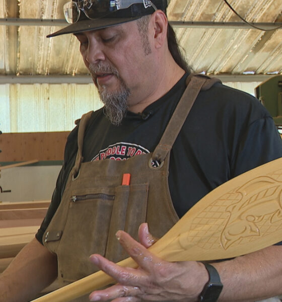 Woodworker in a shop holding a carved wooden surfboard with a fish design