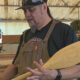 Woodworker in a shop holding a carved wooden surfboard with a fish design