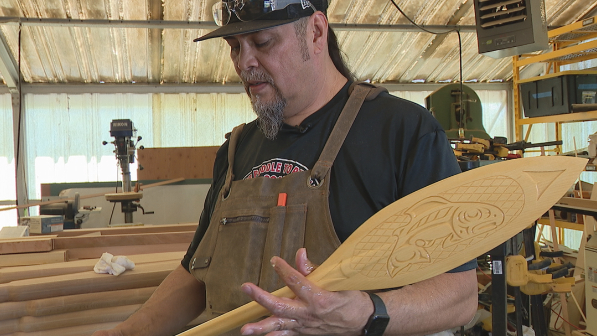 Woodworker in a shop holding a carved wooden surfboard with a fish design