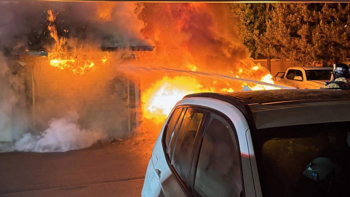 House/garage on fire with bright flames and thick smoke, a white SUV in the foreground watchful bystanders out of frame beyond the blaze