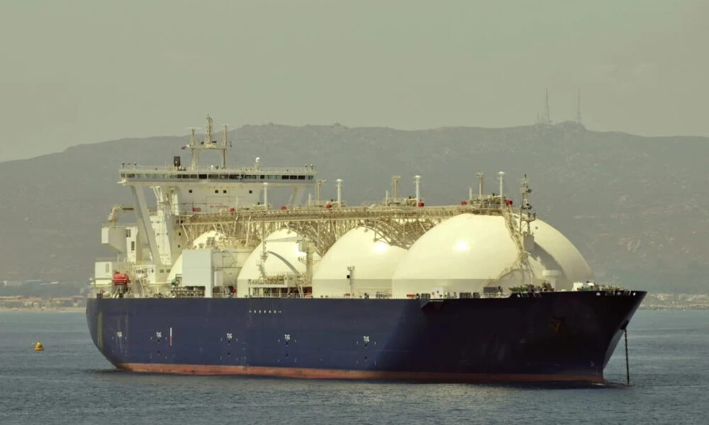 Large LNG carrier ship with white spherical tanks anchored near a coastline, dark hull visible above the water surface.