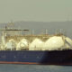 Large LNG carrier ship with white spherical tanks anchored near a coastline, dark hull visible above the water surface.