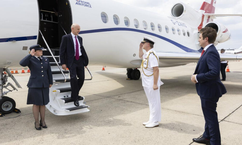 Man in a dark suit steps from a Royal Air Force jet via mobile stairs while a uniformed officer salutes and another suited man watches on the tarmac