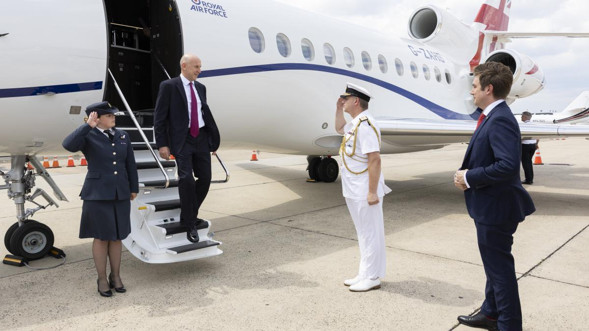Man in a dark suit steps from a Royal Air Force jet via mobile stairs while a uniformed officer salutes and another suited man watches on the tarmac