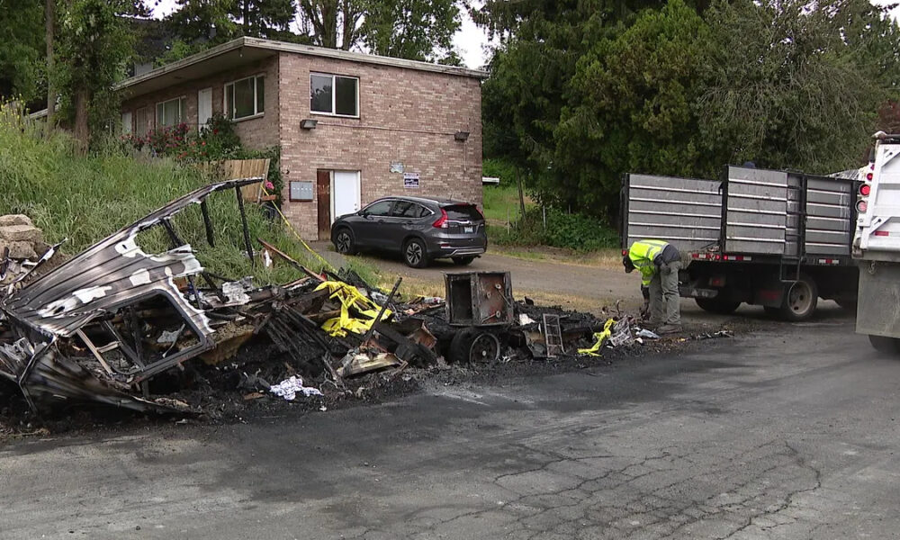 Burnt wreckage along a road with charred debris, a yellow caution tape piece, and a worker in a neon vest inspecting the site near a dump truck and a small office building in the background.