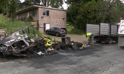 Burnt wreckage along a road with charred debris, a yellow caution tape piece, and a worker in a neon vest inspecting the site near a dump truck and a small office building in the background.