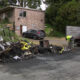 Burnt wreckage along a road with charred debris, a yellow caution tape piece, and a worker in a neon vest inspecting the site near a dump truck and a small office building in the background.
