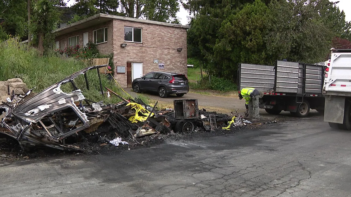 Burnt wreckage along a road with charred debris, a yellow caution tape piece, and a worker in a neon vest inspecting the site near a dump truck and a small office building in the background.