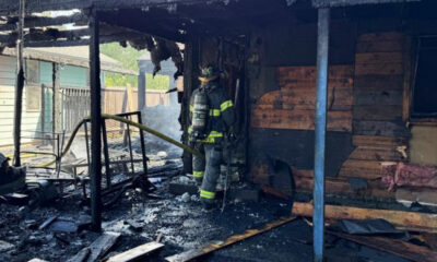 Firefighter in full turnout gear stands amid a burned-out house, holding a hose as charred framing and debris smoulder.