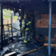 Firefighter in full turnout gear stands amid a burned-out house, holding a hose as charred framing and debris smoulder.