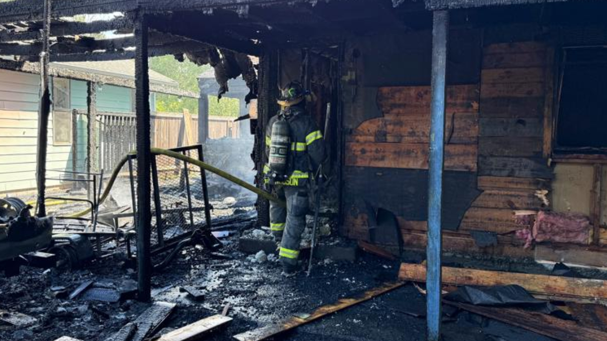 Firefighter in full turnout gear stands amid a burned-out house, holding a hose as charred framing and debris smoulder.