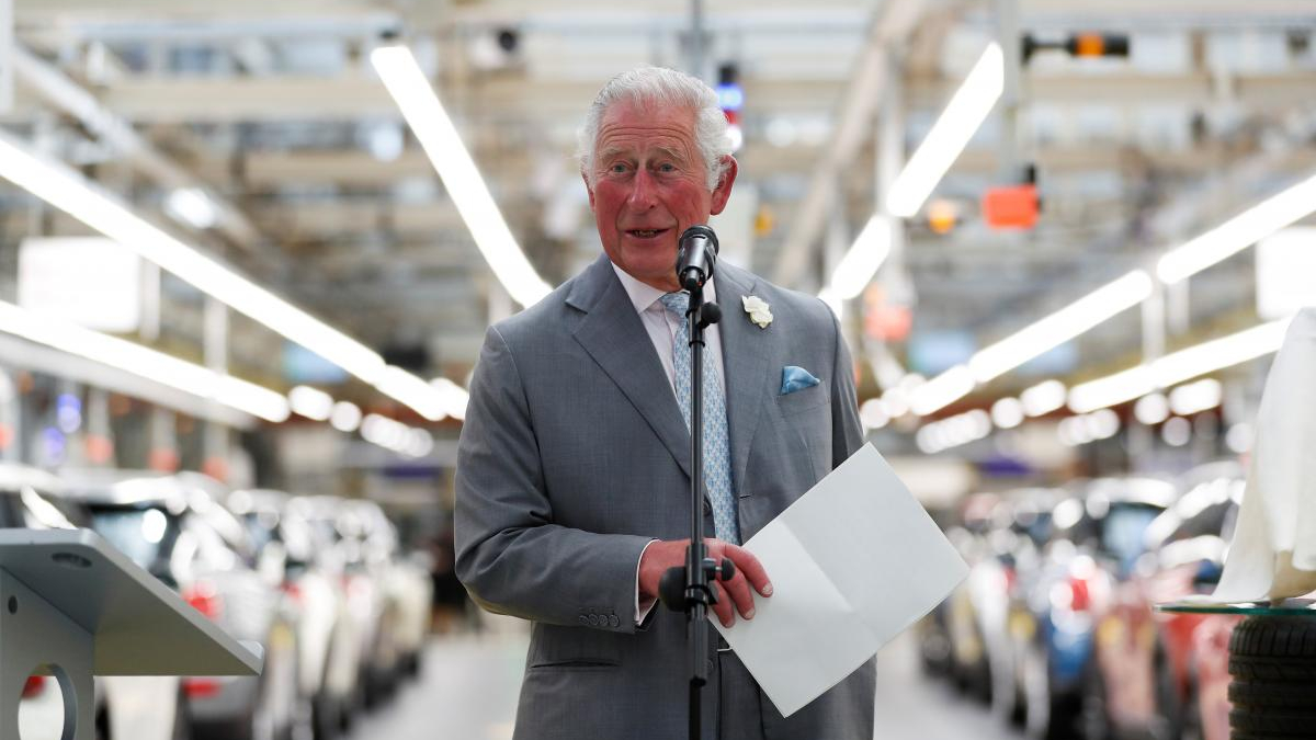 Prince Charles speaks into a microphone at a car manufacturing plant, holding papers.