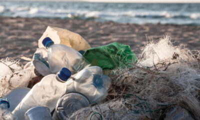 Pile of plastic bottles, cups, and tangled fishing nets littering a sandy beach with the sea in the background.
