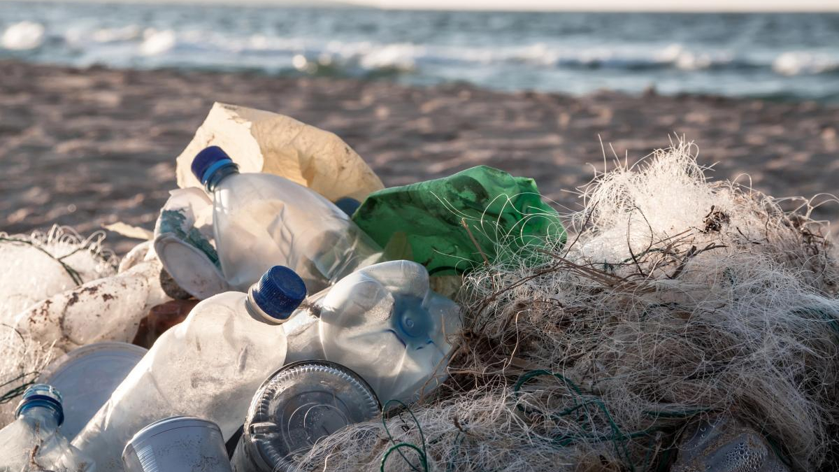 Pile of plastic bottles, cups, and tangled fishing nets littering a sandy beach with the sea in the background.