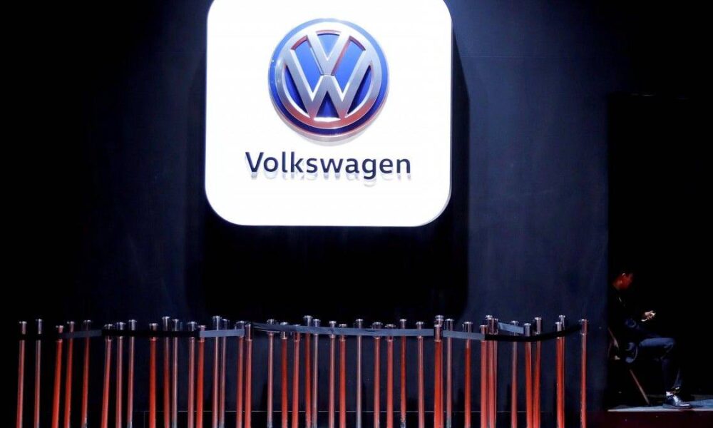 Volkswagen logo on a large illuminated white panel above a row of orange barrier poles in a dim exhibition space, with a person seated to the right.