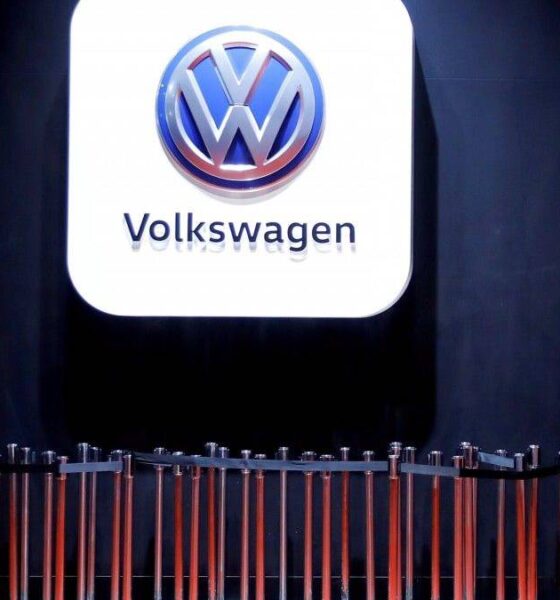 Volkswagen logo on a large illuminated white panel above a row of orange barrier poles in a dim exhibition space, with a person seated to the right.