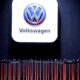 Volkswagen logo on a large illuminated white panel above a row of orange barrier poles in a dim exhibition space, with a person seated to the right.