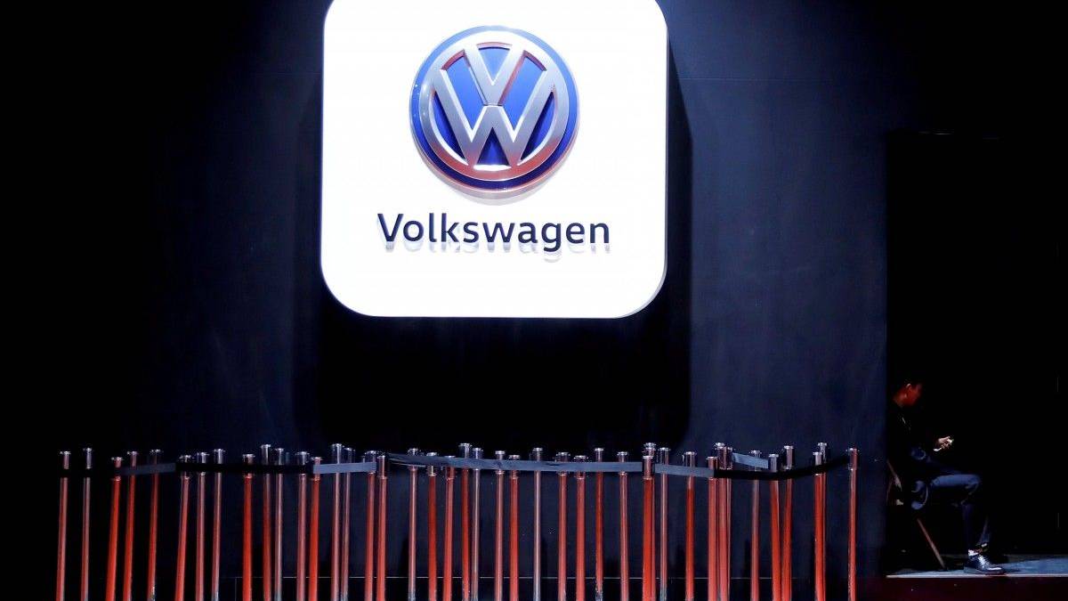 Volkswagen logo on a large illuminated white panel above a row of orange barrier poles in a dim exhibition space, with a person seated to the right.