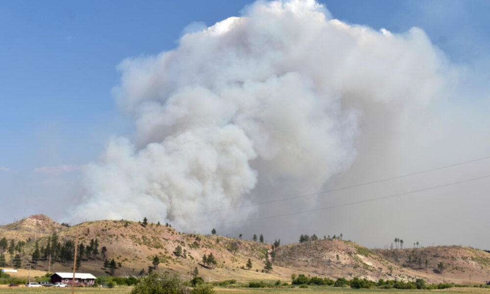 Large plume of white smoke rising from hills near a rural landscape, indicating a wildfire nearby