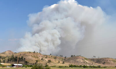 Large plume of white smoke rising from hills near a rural landscape, indicating a wildfire nearby