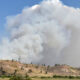 Large plume of white smoke rising from hills near a rural landscape, indicating a wildfire nearby