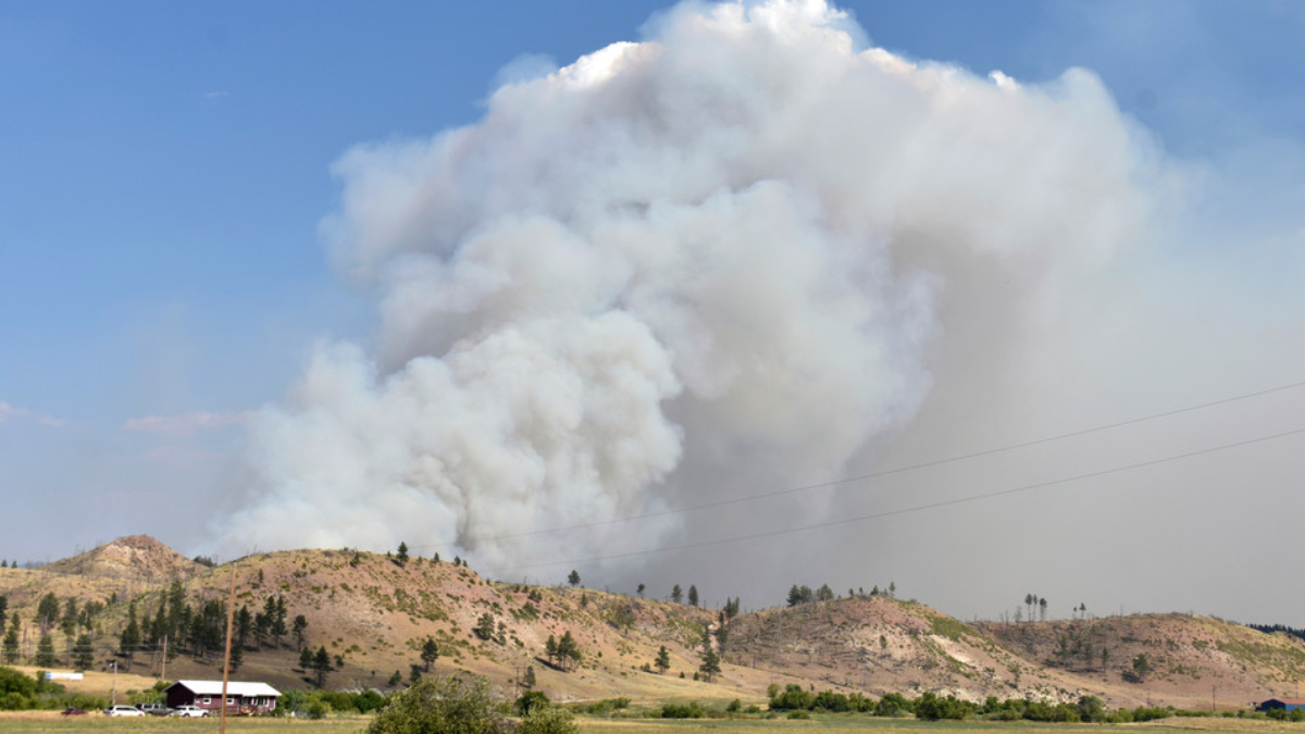 Large plume of white smoke rising from hills near a rural landscape, indicating a wildfire nearby