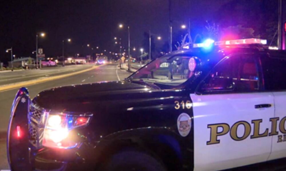 Police SUV with flashing red and blue lights at night, blocking a street during an incident on a dimly lit road down the block.