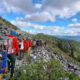 Group of hikers with backpacks walking along a rocky mountain trail overlooking a forested valley under a blue sky with clouds.