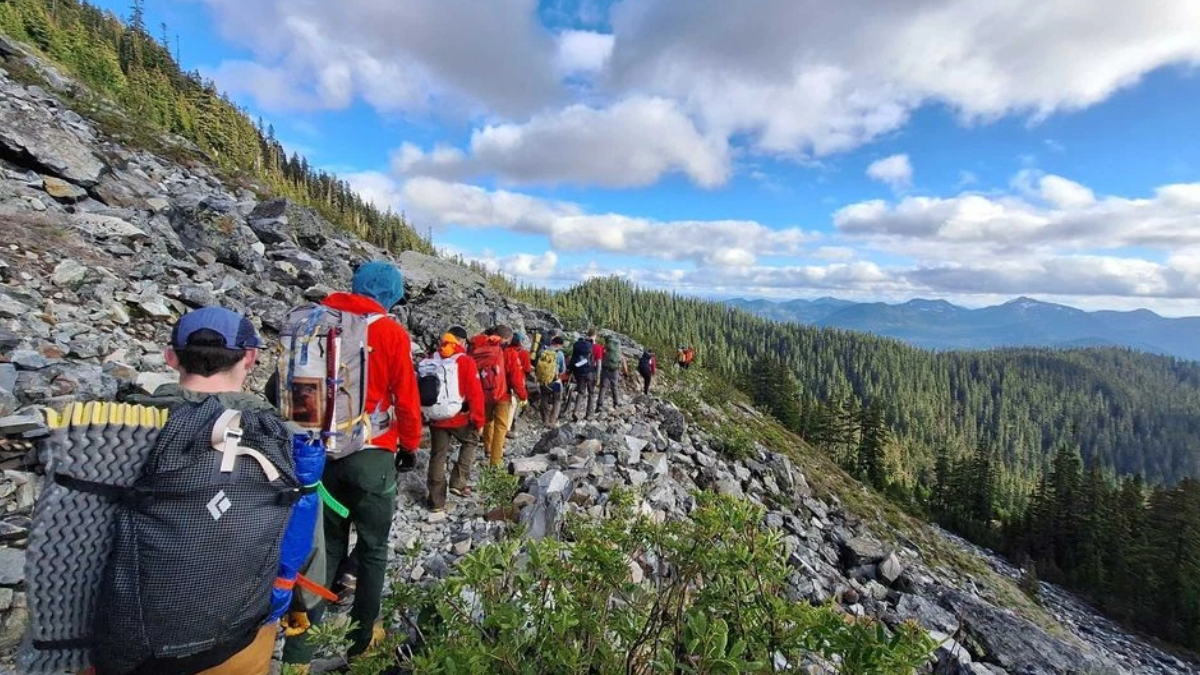 Group of hikers with backpacks walking along a rocky mountain trail overlooking a forested valley under a blue sky with clouds.