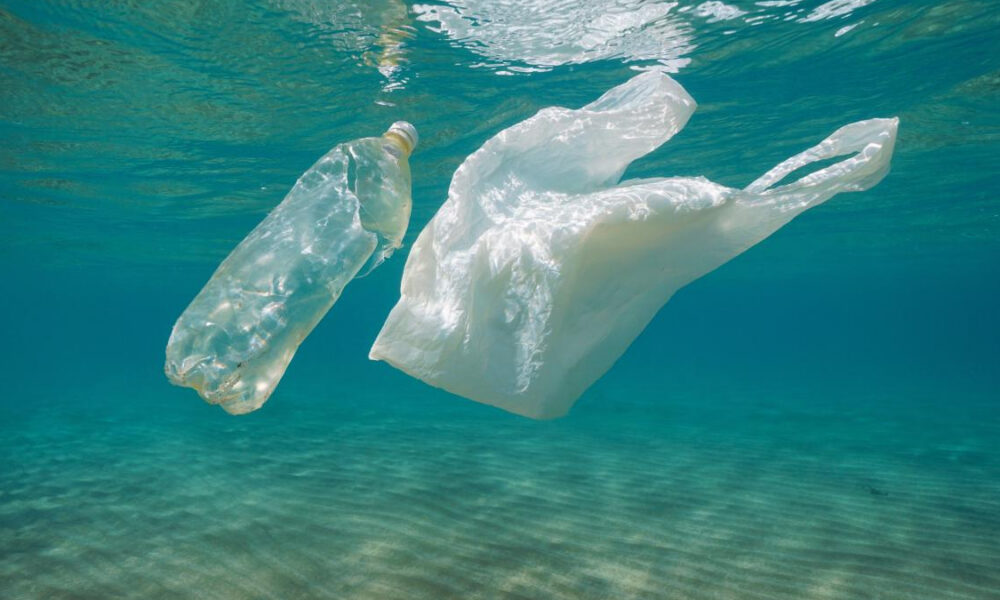 Plastic bottle and plastic bag float underwater in clear blue water, highlighting ocean plastic pollution.