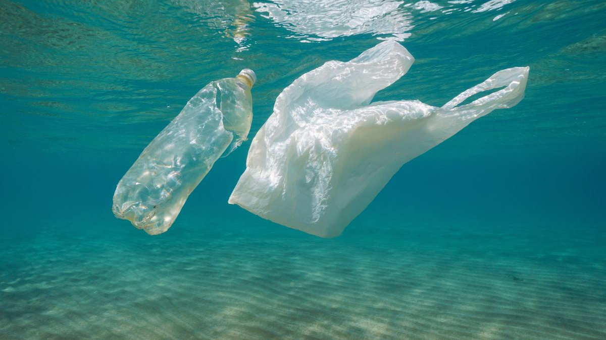 Plastic bottle and plastic bag float underwater in clear blue water, highlighting ocean plastic pollution.