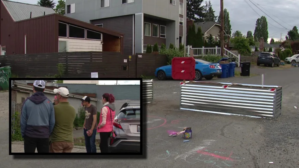 Street scene with a metal barricade blocking the road, chalk drawings on the pavement, a blue car, and several trash bins nearby.