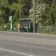 People wait at a green bus shelter on a tree-lined street as a car passes by.