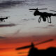 Two quadcopter drones silhouetted against a sunset orange sky, in flight.