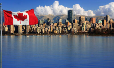 Canadian flag waving on a pole over a blue river, with a city skyline and distant mountains in the background