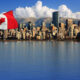 Canadian flag waving on a pole over a blue river, with a city skyline and distant mountains in the background