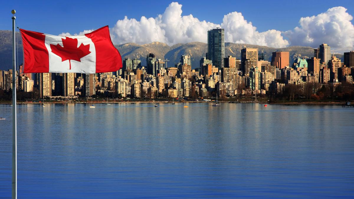 Canadian flag waving on a pole over a blue river, with a city skyline and distant mountains in the background