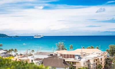 Coastal town with beige rooftops, palm trees, and a turquoise sea; a cruise ship sits offshore in the distance.
