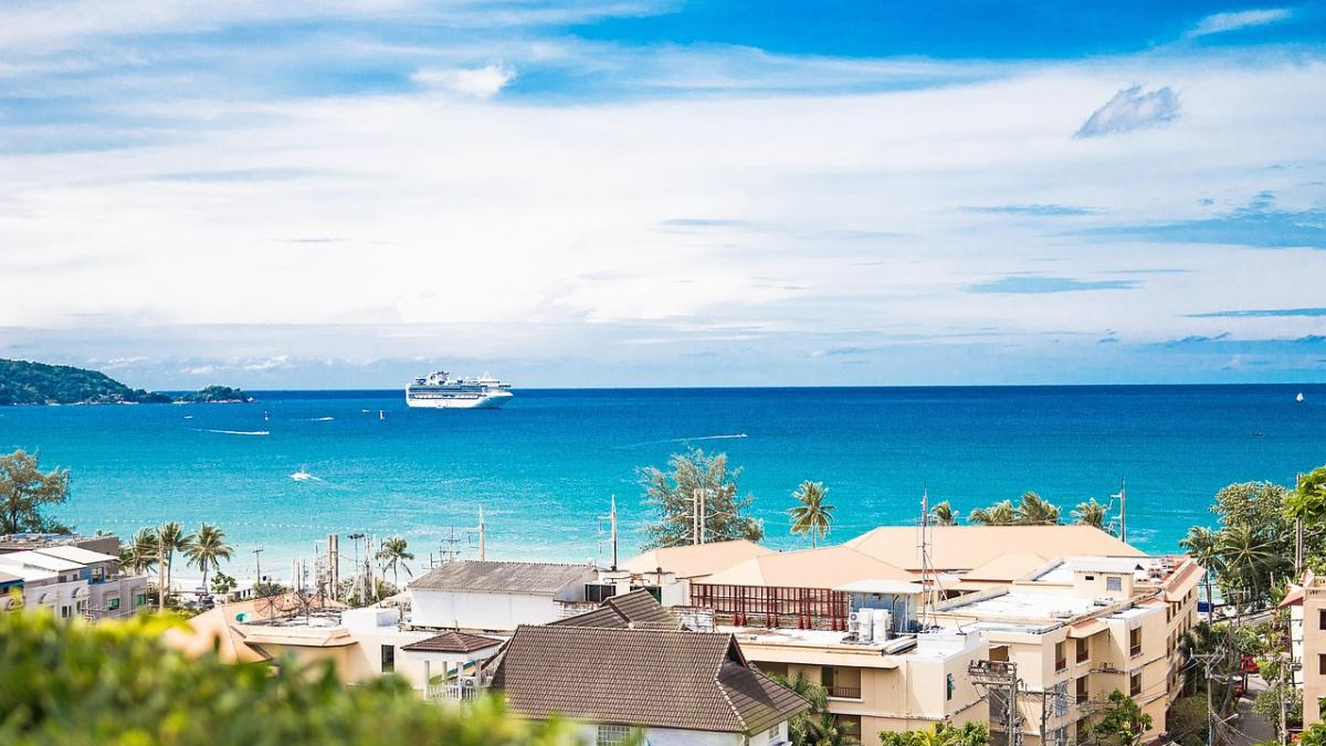 Coastal town with beige rooftops, palm trees, and a turquoise sea; a cruise ship sits offshore in the distance.